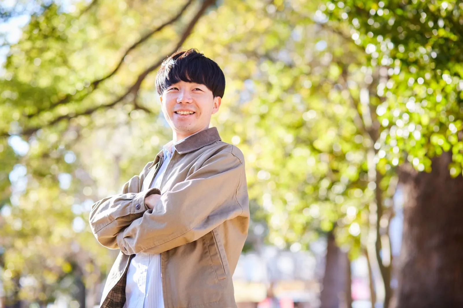 Tak smiling with his arms crossed under green trees