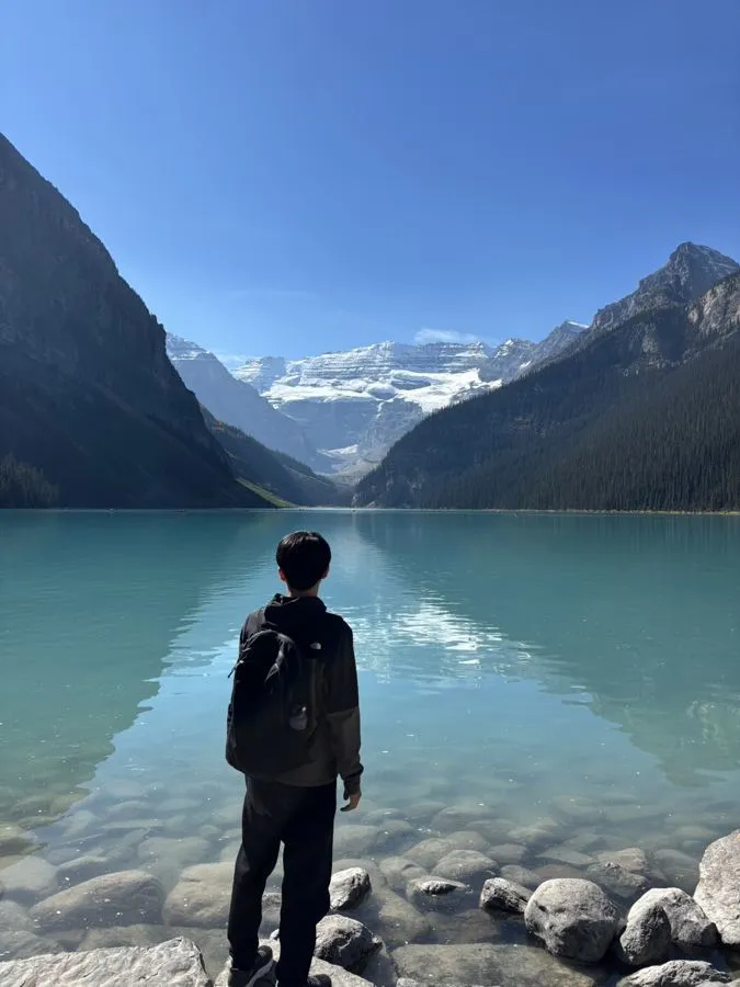 Tak from behind, standing on rocks by a turquoise lake with snowy mountains